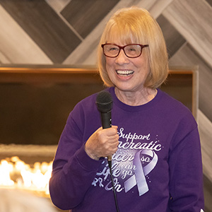 Woman in purple cancer shirt and red framed glasses hold a microphone smiling.