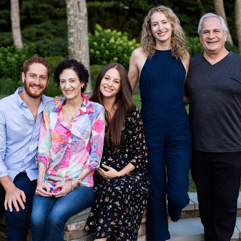 Five adults pose for a family photo with lush green grass and bushes behind them. The three on the left sit while the two on the right stand.