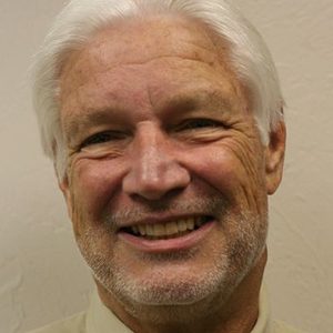 Man with white hair smiles for the camera wearing a beige button up shirt.