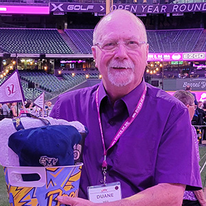 Man wearing a purple button up and purple nametag lanyard is standing inside a ballpark hold a prize basket.